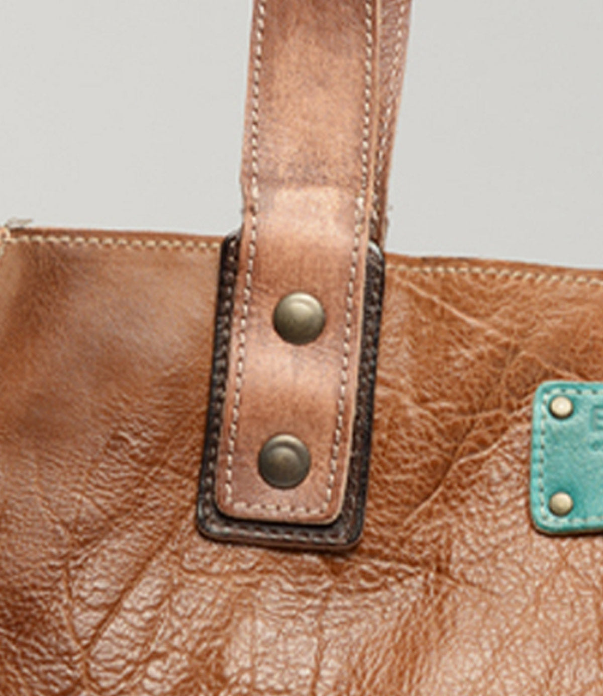 Close-up of the Studio Strap Underlay by Bed Stu, featuring a brown leather strap adorned with two metal rivets. A green leather patch appears in the bottom right corner, emphasizing the unique design of this personalized bag accessory.