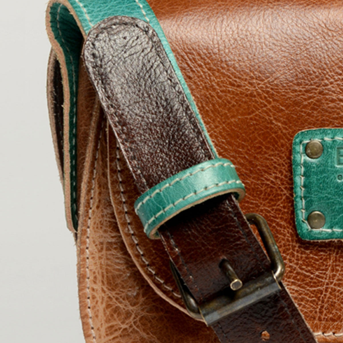 Close-up of the Studio Buckle Strap by Bed Stu, featuring a brown leather bag with a teal strap and teal patch showcasing visible stitching and a metal buckle—perfect for expressing your own style with this personalized bag.