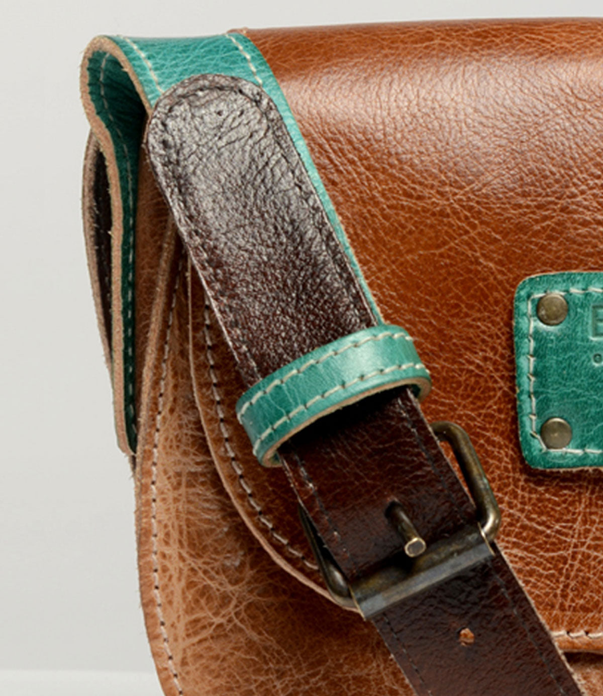 Close-up of the Studio Buckle Strap by Bed Stu, featuring a brown leather bag with a teal strap and teal patch showcasing visible stitching and a metal buckle—perfect for expressing your own style with this personalized bag.