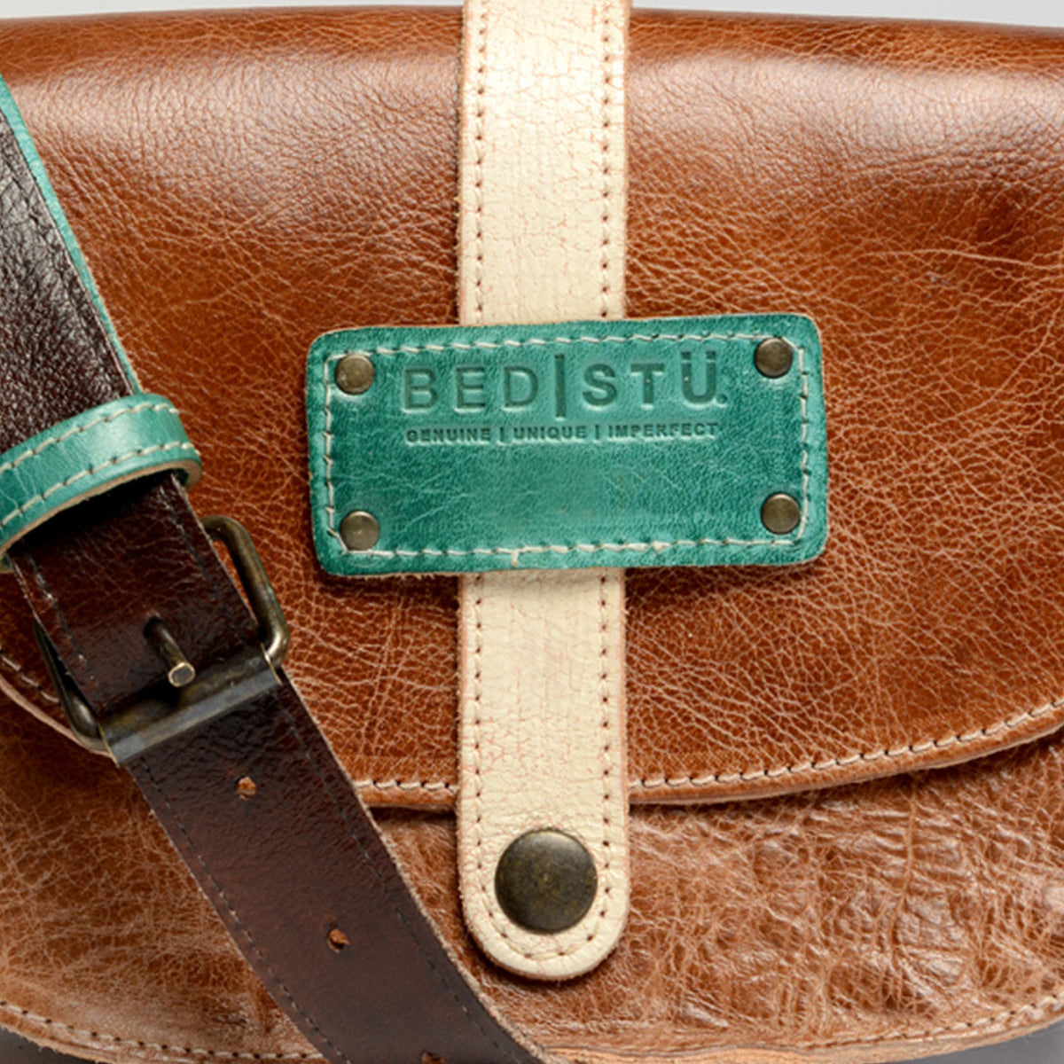 The Studio Snap Strap is a brown leather bag featuring a contrasting light tan strap and buckle. It has a turquoise label from Bed Stu that reads "Bed Stu" with the words "Genuine Unique Imperfect" below it, making it perfect to reflect your own style.