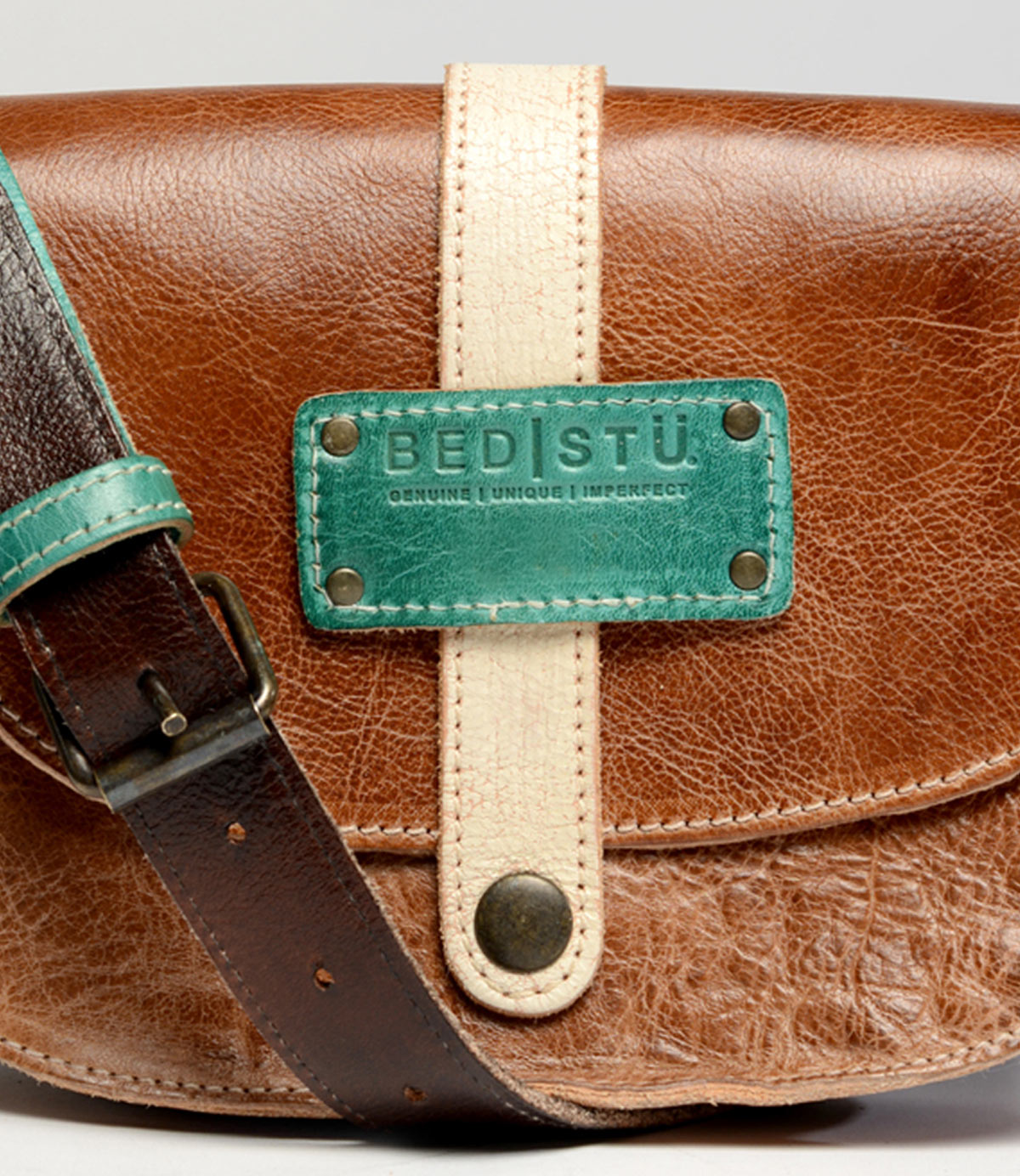 The Studio Snap Strap is a brown leather bag featuring a contrasting light tan strap and buckle. It has a turquoise label from Bed Stu that reads "Bed Stu" with the words "Genuine Unique Imperfect" below it, making it perfect to reflect your own style.