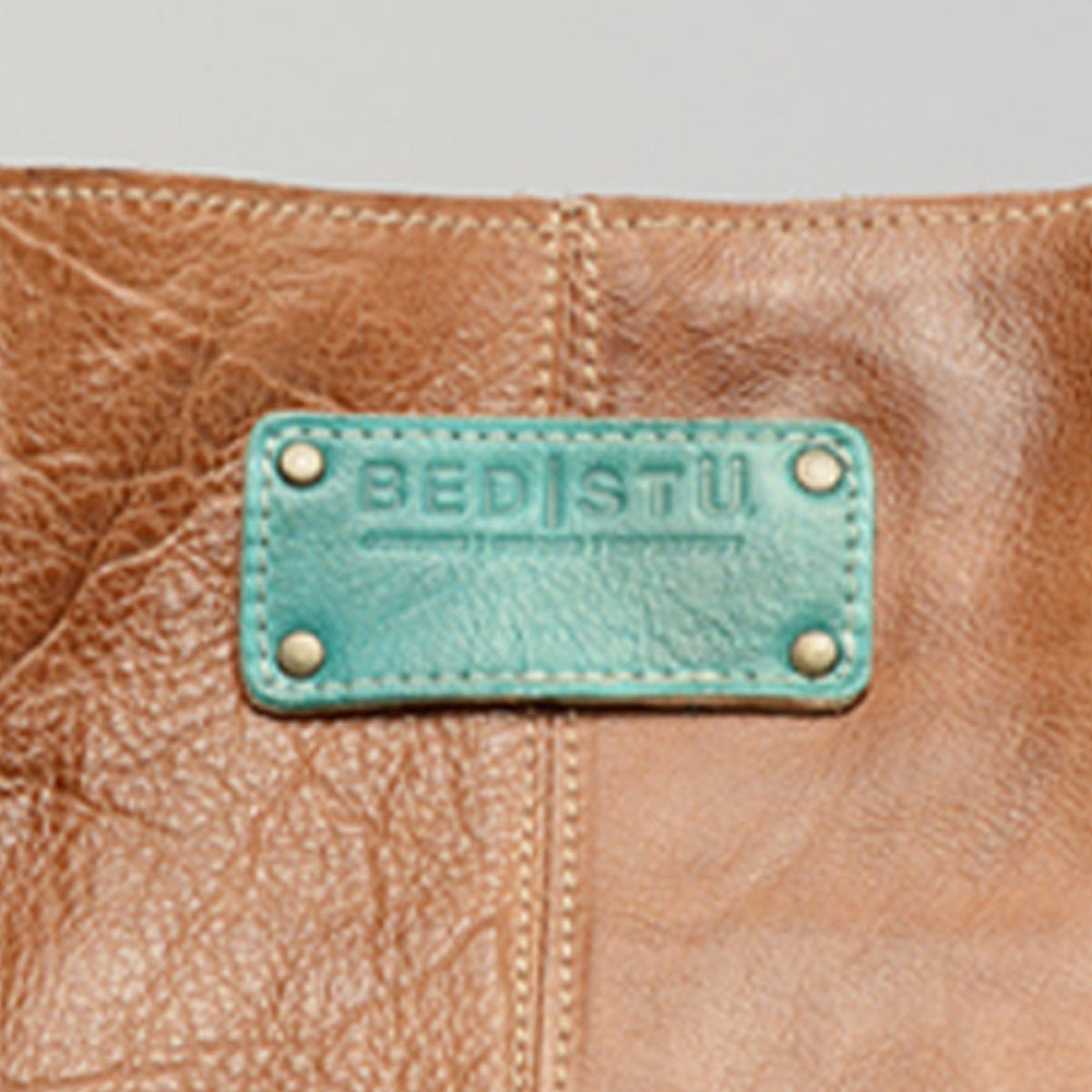 Close-up of a brown leather item with a teal patch featuring the "Studio Logo Patch" by Bed Stu, showcasing the unique colors and combinations perfect for a personalized bag.