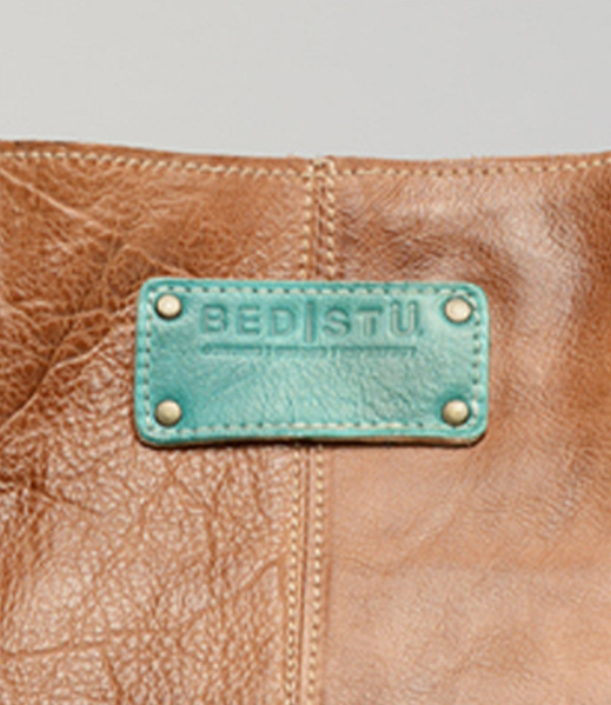 Close-up of a brown leather item with a teal patch featuring the "Studio Logo Patch" by Bed Stu, showcasing the unique colors and combinations perfect for a personalized bag.
