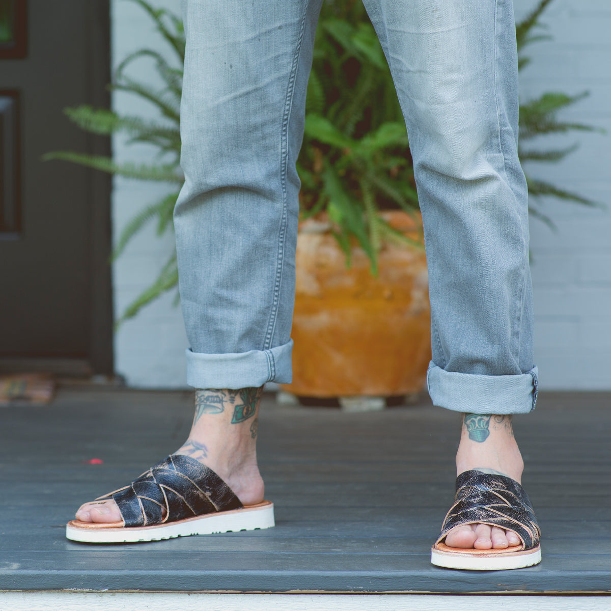 A person wearing Abraham Light jeans and Bed Stu black leather sandals stands on a white doorstep, revealing tattooed ankles.