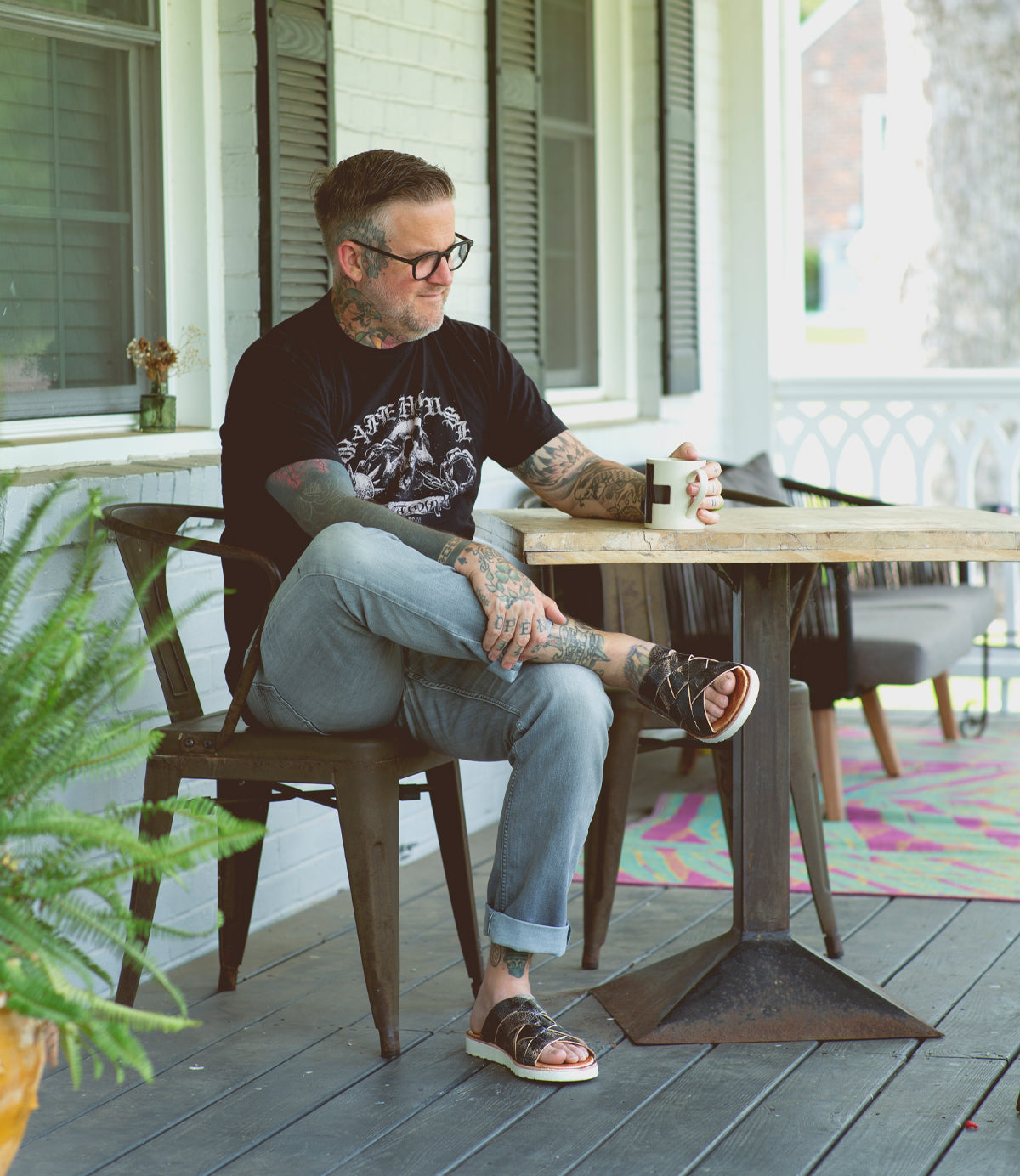 A tattooed man wearing glasses, a black t-shirt, and gray pants sits at a wooden table on a porch, holding an Abraham Light mug and wearing Bed Stu leather sandals.