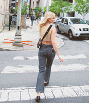 A woman with blonde hair walks across a crosswalk on a city street, wearing a beige crop top, jeans, and black shoes. She is holding a small item in her right hand and has a Bed Stu Abundance sling bag over her shoulder, allowing for hands-free convenience.