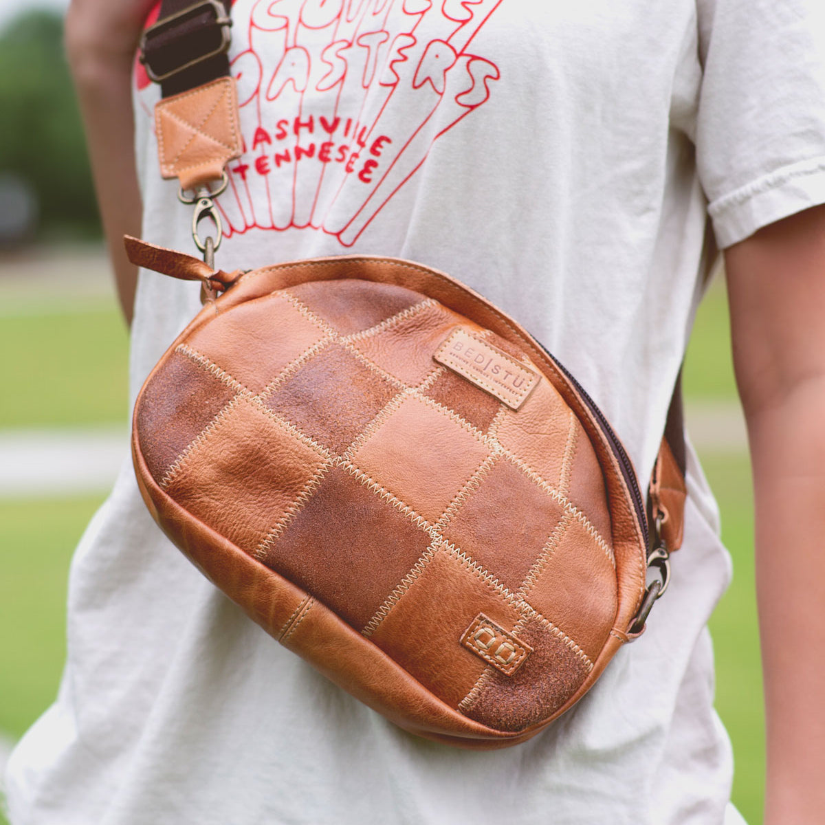 A person wearing a white T-shirt with a graphic, carrying an Abundance sling bag by Bed Stu in a brown patchwork leather design.