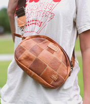 A person wearing a white T-shirt with a graphic, carrying an Abundance sling bag by Bed Stu in a brown patchwork leather design.