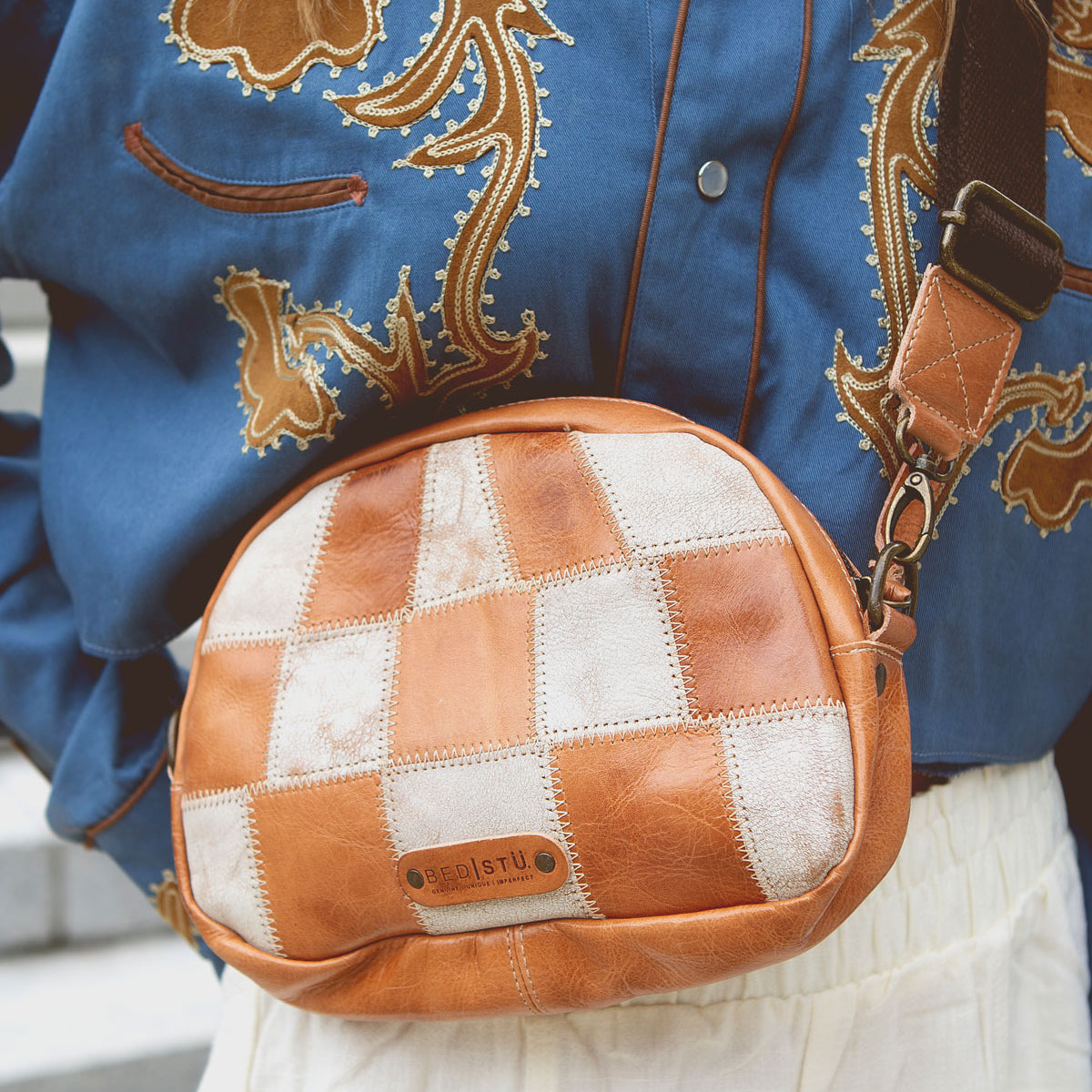 Person wearing a blue shirt with an ornate design and a Bed Stu Abundance sling bag with a checkered pattern, in shades of brown and white, perfect for hands-free convenience.