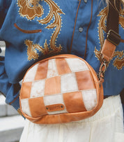 Person wearing a blue shirt with an ornate design and a Bed Stu Abundance sling bag with a checkered pattern, in shades of brown and white, perfect for hands-free convenience.