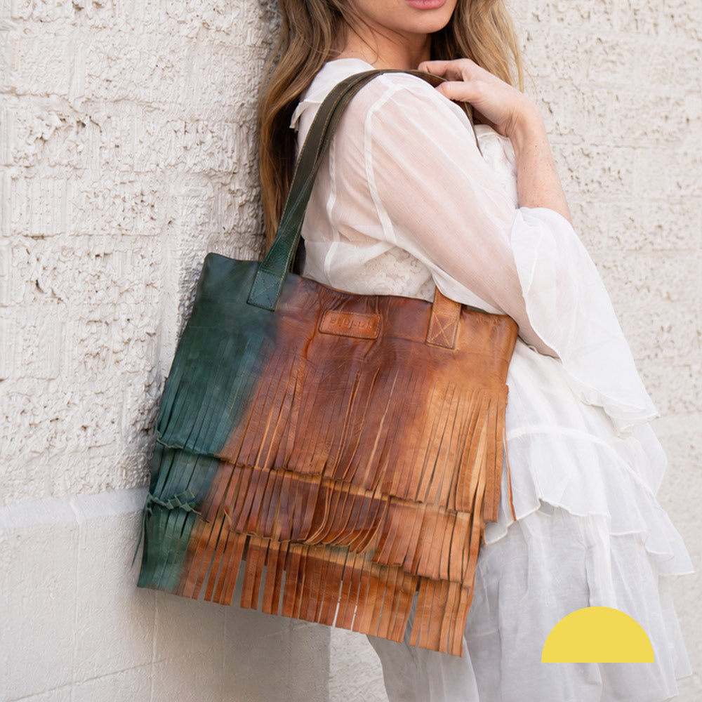 Person in a flowing white dress carries a large brown leather tote with dark green side panels and a textured, fringed design.