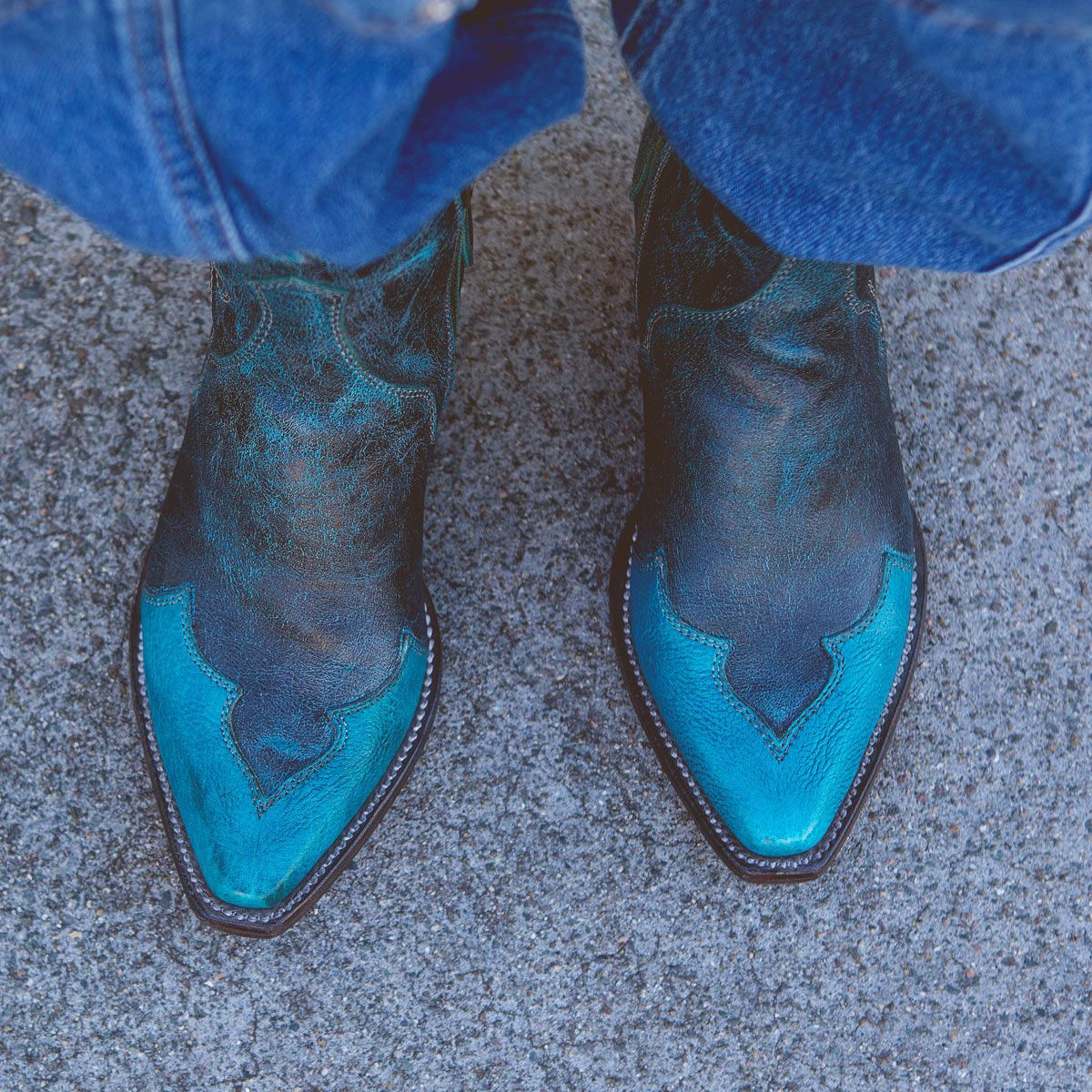 A pair of worn blue and black Ace cowboy boots by Bed Stu, crafted from full-grain vegetable-tanned leather, rest on a rough grey concrete surface, with the bottom of blue jeans visible. This touch of modern western style blends seamlessly with their rugged backdrop.