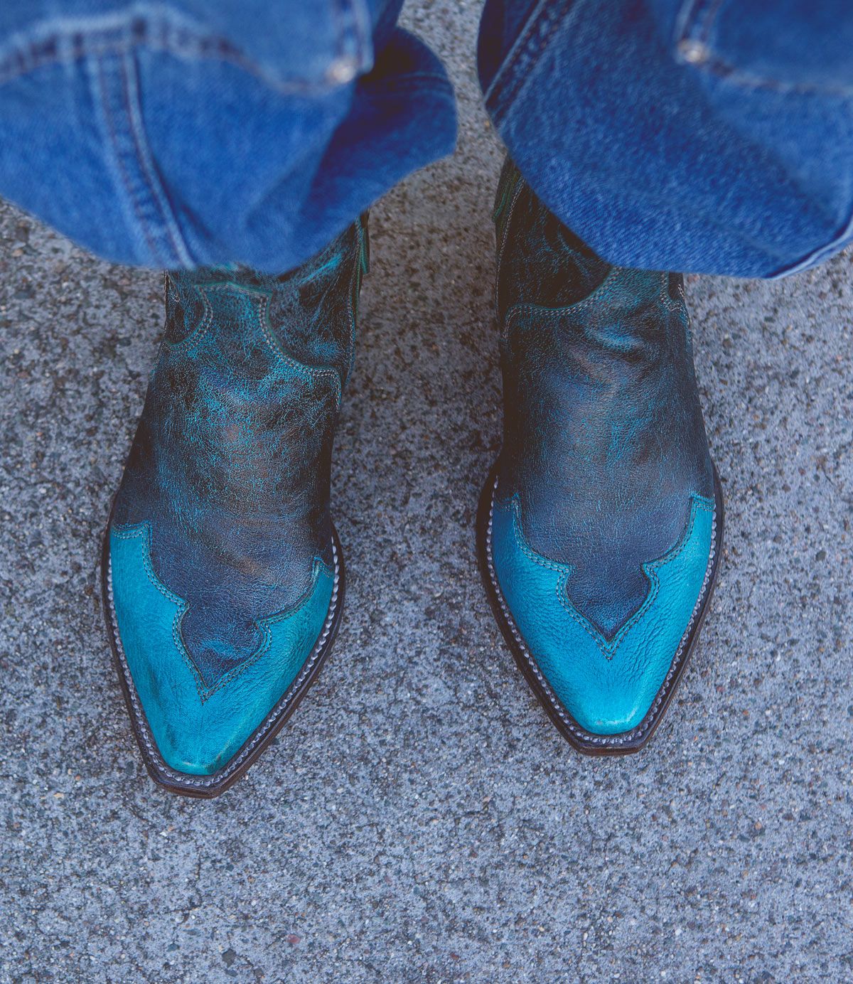 A pair of worn blue and black Ace cowboy boots by Bed Stu, crafted from full-grain vegetable-tanned leather, rest on a rough grey concrete surface, with the bottom of blue jeans visible. This touch of modern western style blends seamlessly with their rugged backdrop.