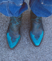 A pair of worn blue and black Ace cowboy boots by Bed Stu, crafted from full-grain vegetable-tanned leather, rest on a rough grey concrete surface, with the bottom of blue jeans visible. This touch of modern western style blends seamlessly with their rugged backdrop.