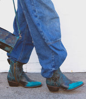 A person in blue jeans is walking, sporting Ace teal and brown ankle boots crafted from full-grain vegetable-tanned leather by Bed Stu, and carrying a matching handbag.