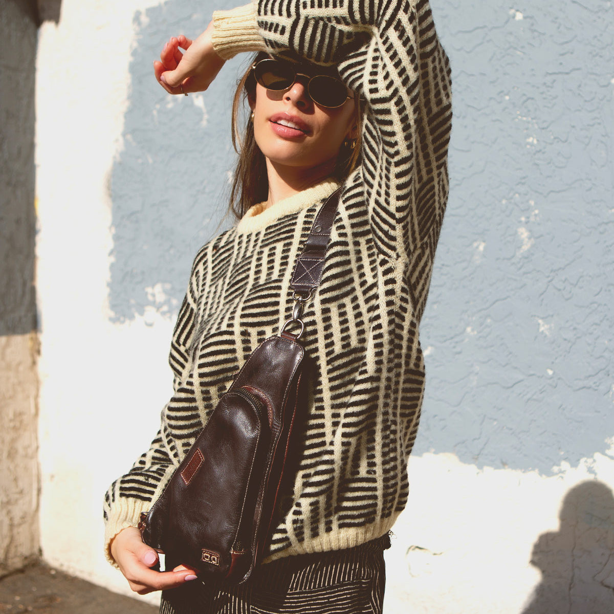 A person wearing a geometric-patterned sweater, sunglasses, and the Bed Stu Adept compact sling bag made from vegetable-tanned leather poses with one arm raised against a textured wall background.