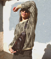 A person wearing a geometric-patterned sweater, sunglasses, and the Bed Stu Adept compact sling bag made from vegetable-tanned leather poses with one arm raised against a textured wall background.