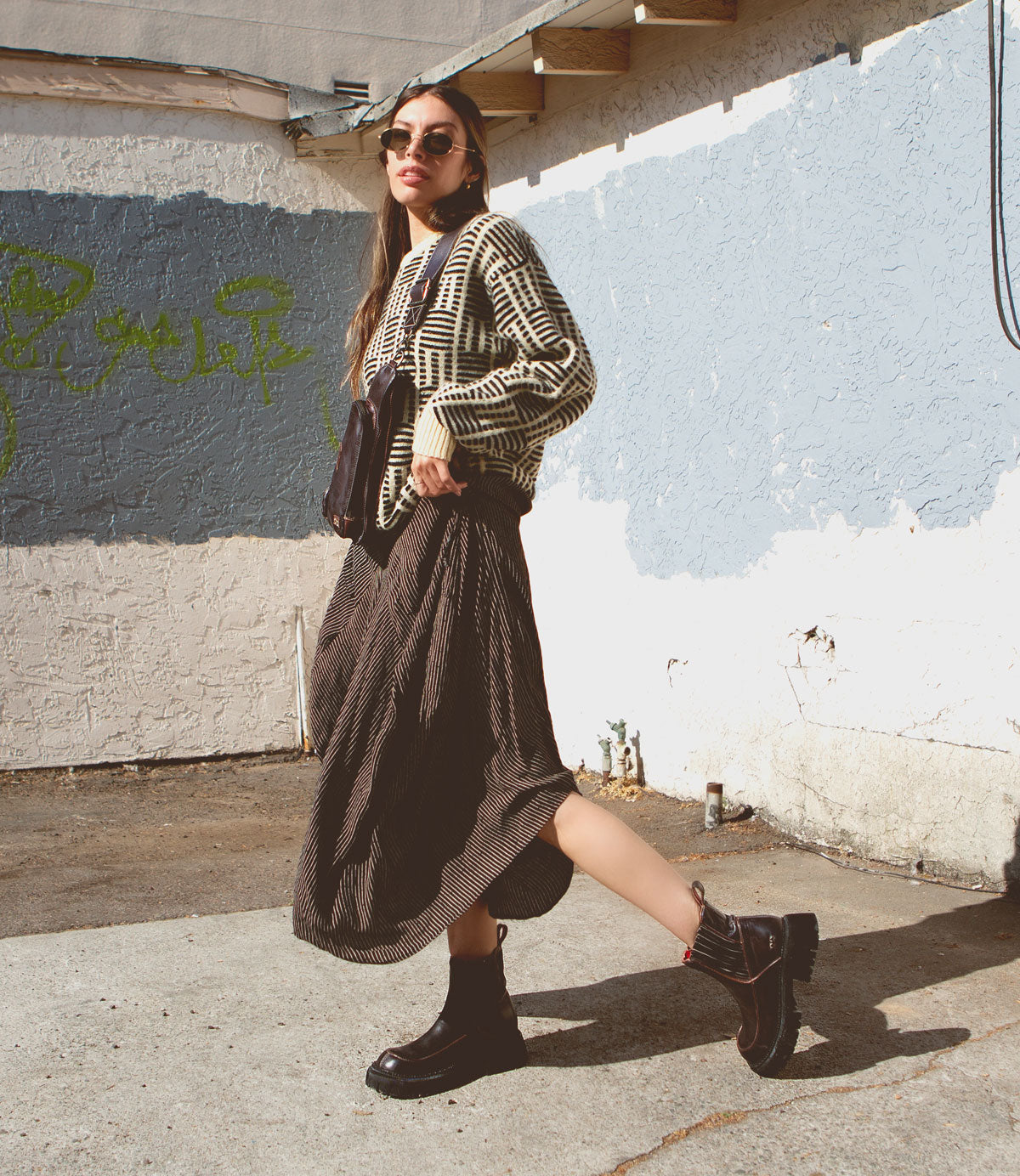 A woman with long hair, wearing sunglasses, a patterned cardigan, a flowing skirt, and black boots, stands and poses in an urban alleyway with graffiti and blue textured walls. She carries the compact **Adept** sling bag from **Bed Stu**, made of vegetable-tanned leather—a perfect travel companion for her city adventures.