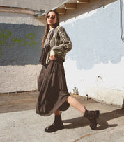 A woman with long hair, wearing sunglasses, a patterned cardigan, a flowing skirt, and black boots, stands and poses in an urban alleyway with graffiti and blue textured walls. She carries the compact **Adept** sling bag from **Bed Stu**, made of vegetable-tanned leather—a perfect travel companion for her city adventures.