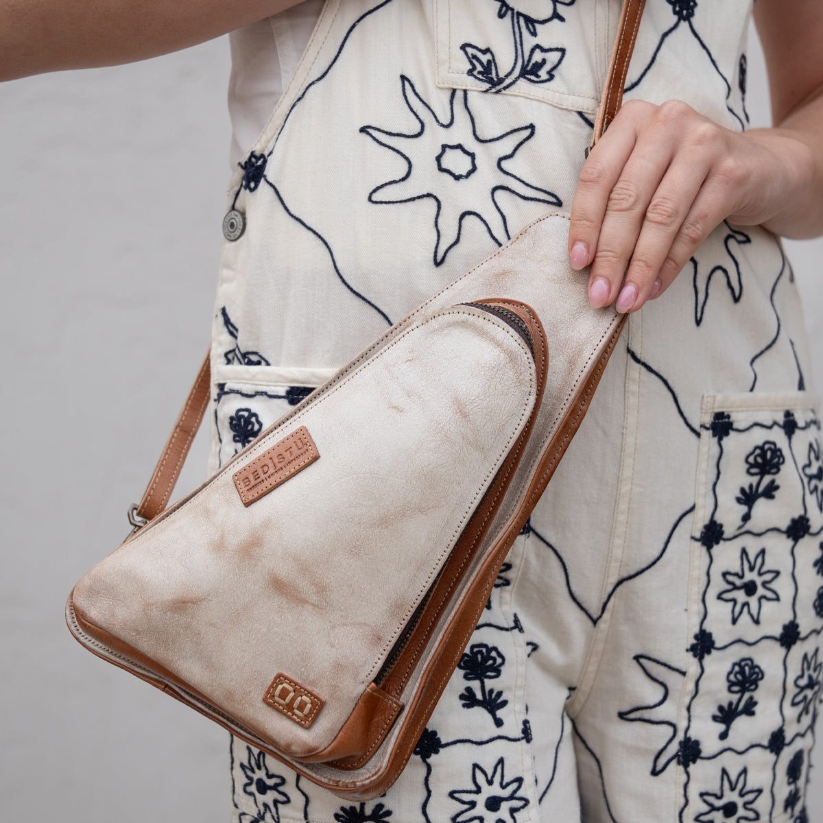 A person in cream overalls with blue floral embroidery holds the "Adept" crossbody bag by Bed Stu, a beige triangular accessory made from vegetable tanned leather with brown trim and branded patches.