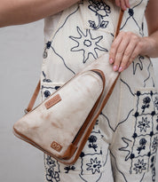 A person in cream overalls with blue floral embroidery holds the "Adept" crossbody bag by Bed Stu, a beige triangular accessory made from vegetable tanned leather with brown trim and branded patches.