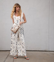 A woman in a white patterned jumpsuit, sandals, and the Bed Stu Adept vegetable tanned leather crossbody bag stands on a sidewalk against a plain, textured wall.