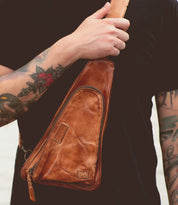 Close-up of a person with tattoos wearing a black shirt, holding the Bed Stu Adept, a versatile crossbody bag made from vegetable tanned leather with a single strap across their chest.