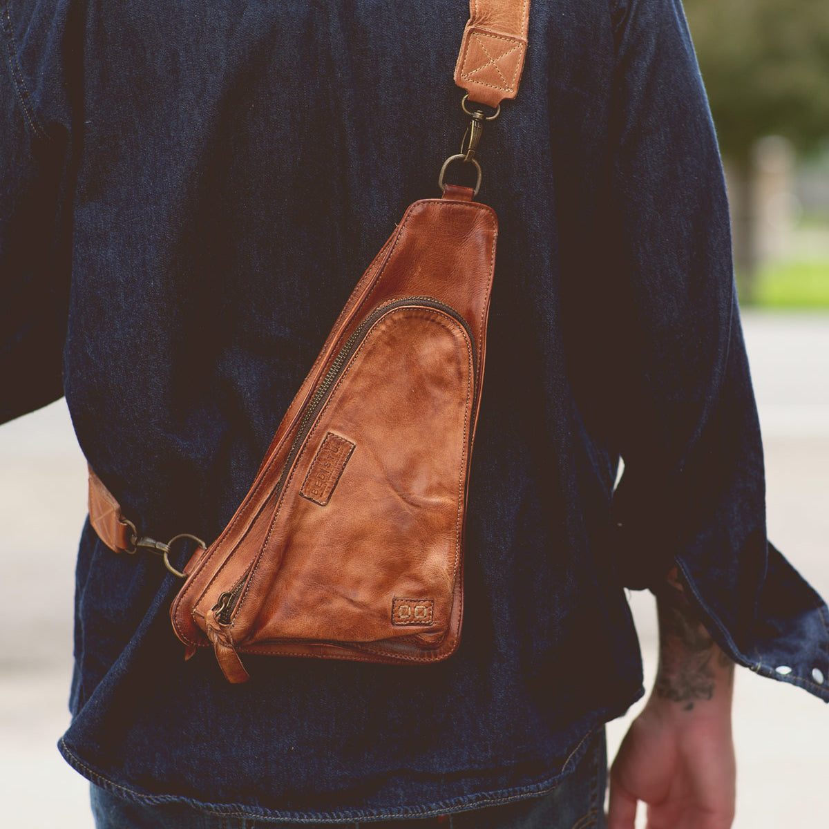 A person wearing a blue denim shirt and jeans is carrying a brown vegetable tanned leather sling bag with a triangular shape, featuring a main zipper compartment and an adjustable shoulder strap. The Adept by Bed Stu makes for the perfect travel companion.
