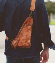 A person wearing a blue denim shirt and jeans is carrying a brown vegetable tanned leather sling bag with a triangular shape, featuring a main zipper compartment and an adjustable shoulder strap. The Adept by Bed Stu makes for the perfect travel companion.
