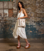 A woman in a white sleeveless dress and platform sandals stands indoors with the Bed Stu Aiken convertible crossbody bag, which features an adjustable strap, in front of a textured brick and plaster wall.