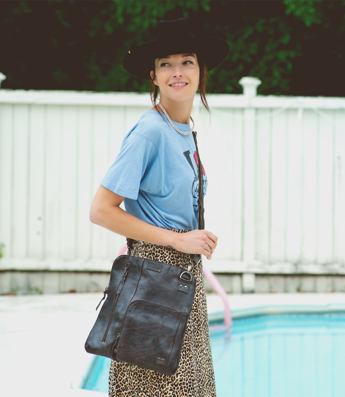 A woman in a hat and leopard skirt standing next to a pool, confidently carrying the Bed Stu Aiken, a convertible crossbody bag made of vegetable-tanned leather.