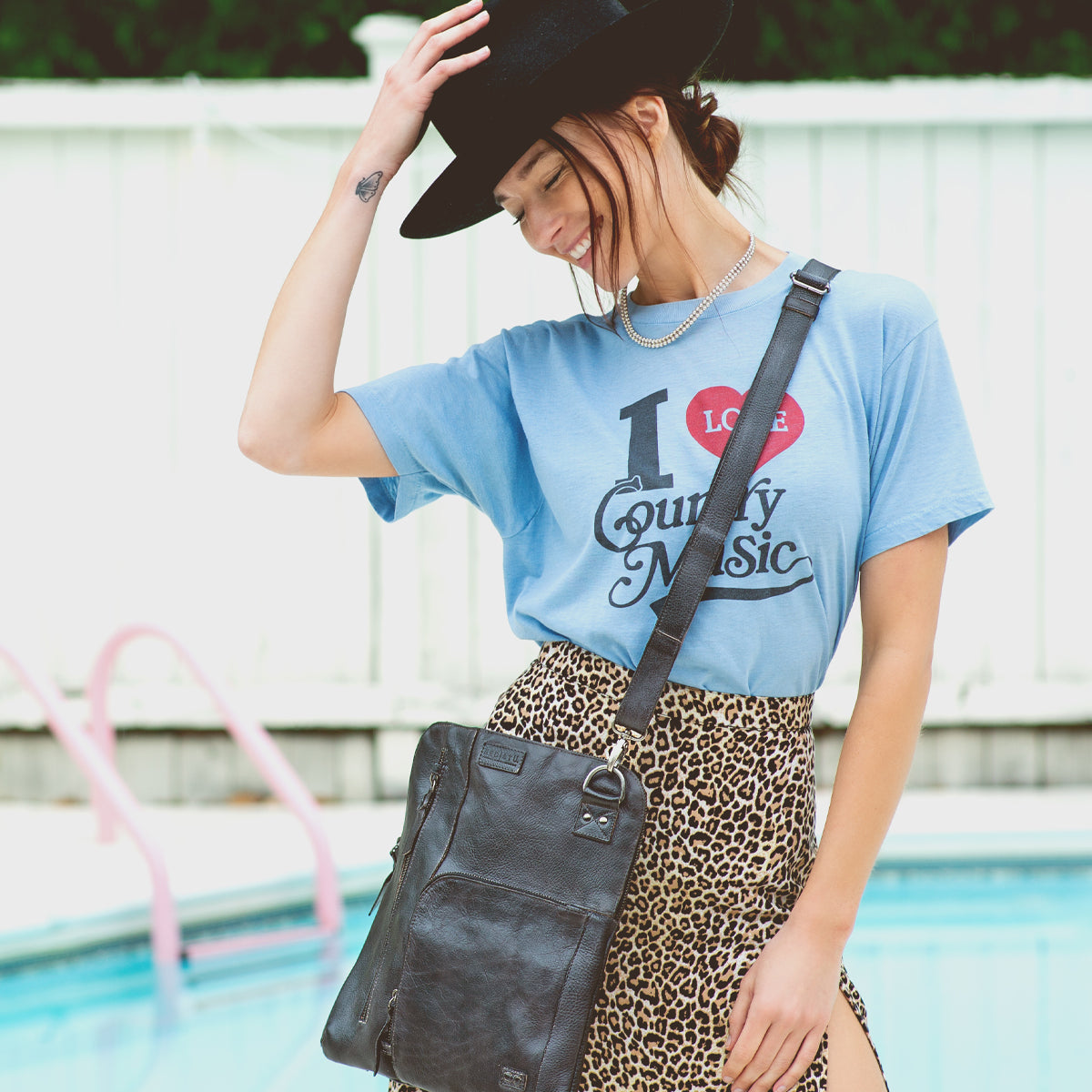 A woman wearing a t-shirt and leopard skirt standing next to a pool, holding a Bed Stu Aiken crossbody bag.