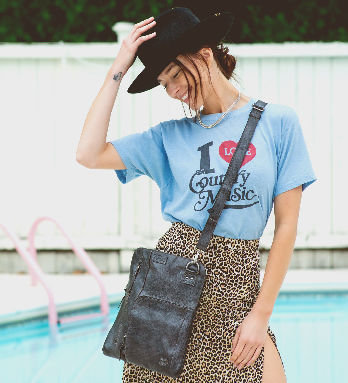 By the pool, a woman smiles with her hand on her hat, wearing a black hat, blue "I Love Country Music" shirt, leopard print skirt, and the Bed|Stü Aiken crossbody bag crafted from full-grain leather with an adjustable strap.