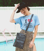 A woman wearing a t-shirt and leopard skirt standing next to a pool, holding a Bed Stu Aiken crossbody bag.