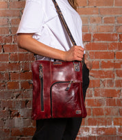 A person wearing a white tee and black pants holds the Bed Stu Aiken, a red leather convertible crossbody with zippers, pockets, and an adjustable strap, while standing in front of a brick wall.
