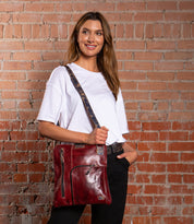 A woman stands against a brick wall, wearing a white tee and black pants, carrying the Bed Stu Aiken red leather convertible crossbody bag with zippered pockets and an adjustable strap.