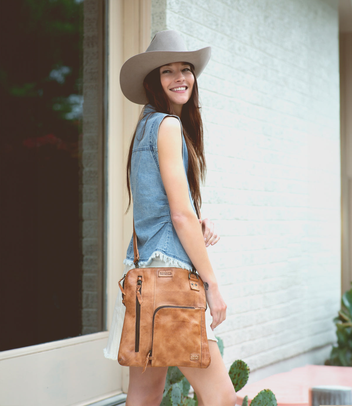 A woman wearing a cowboy hat and shorts is holding a Bed Stu Aiken leather crossbody bag.