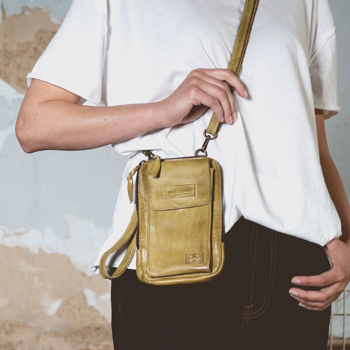 A person in a white t-shirt and jeans is holding Bed Stu's Alelike, a small yellow crossbody phone holder, ideal for everyday functionality.