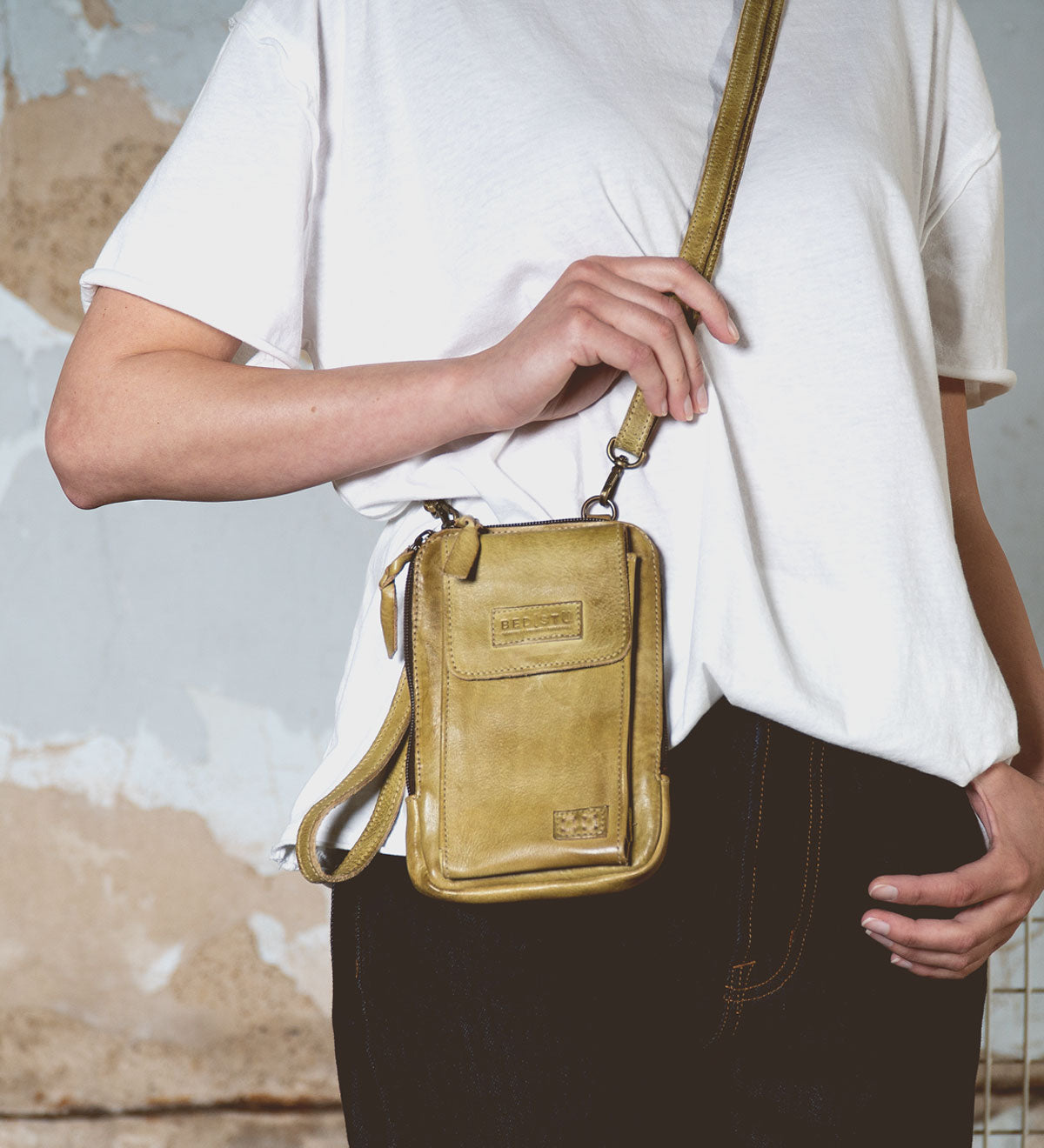 A person in a white t-shirt and jeans is holding Bed Stu's Alelike, a small yellow crossbody phone holder, ideal for everyday functionality.
