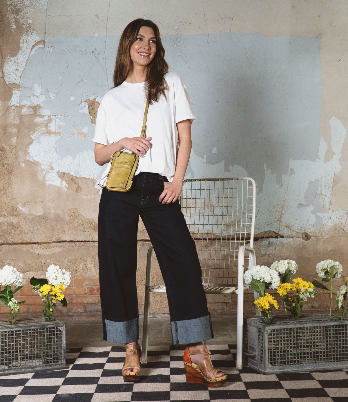 A woman wearing a white shirt and dark jeans stands beside a chair on a checkered floor, surrounded by flower crates and against a worn wall. She carries the Bed Stu Alelike, a stylish crossbody phone holder crafted from premium leather.