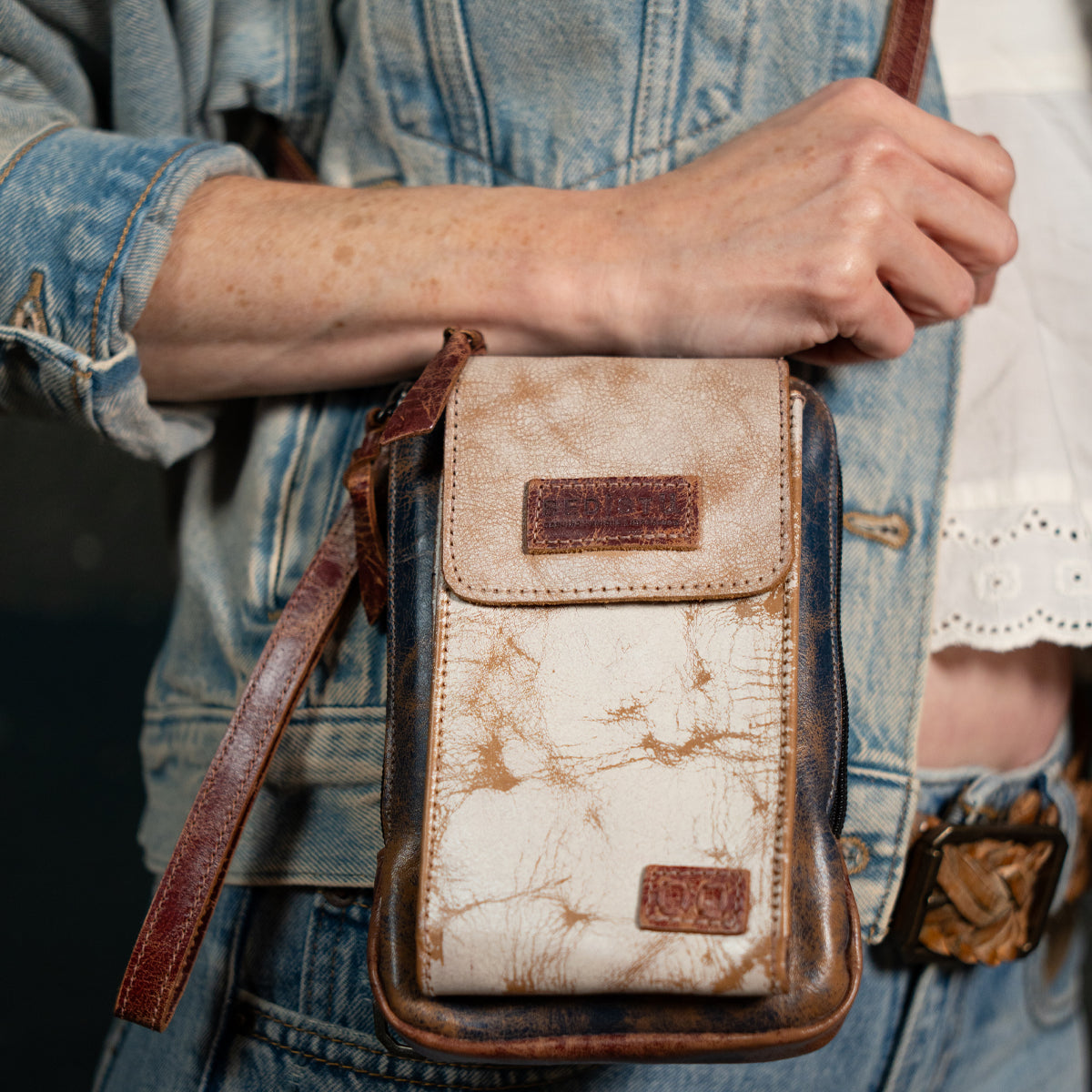 Wearing a denim jacket and belt, a person carries the Bed Stu Alelike—an elegant compact leather crossbody bag with a marbled pattern and brown strap—ideal for keeping travel essentials close at hand.