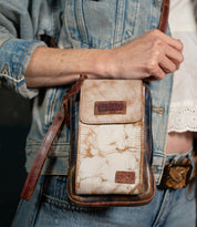 Wearing a denim jacket and belt, a person carries the Bed Stu Alelike—an elegant compact leather crossbody bag with a marbled pattern and brown strap—ideal for keeping travel essentials close at hand.