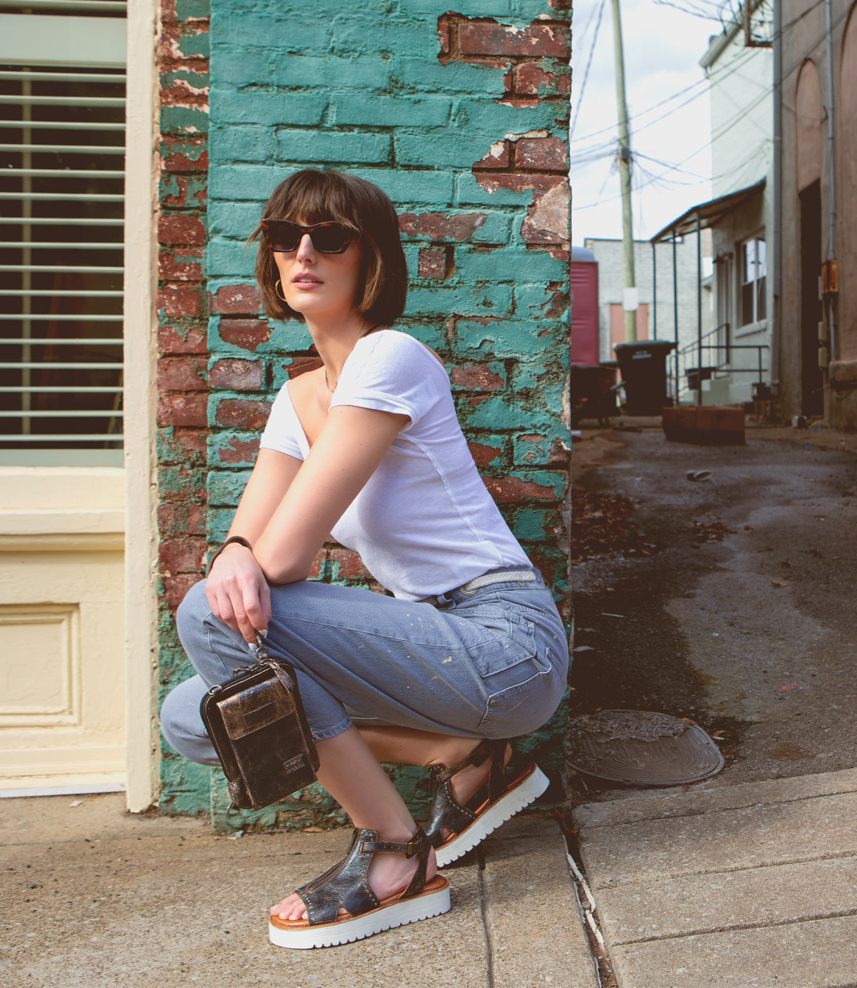 A woman crouching down next to a building, sporting a Bed Stu Alelike premium leather crossbody phone holder.