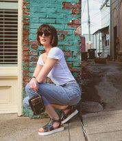 A woman crouching down next to a building, sporting a Bed Stu Alelike premium leather crossbody phone holder.
