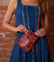 Against a brick wall background, a woman in a blue denim dress holds the Bed Stu Alelike, a compact, premium leather crossbody phone holder.