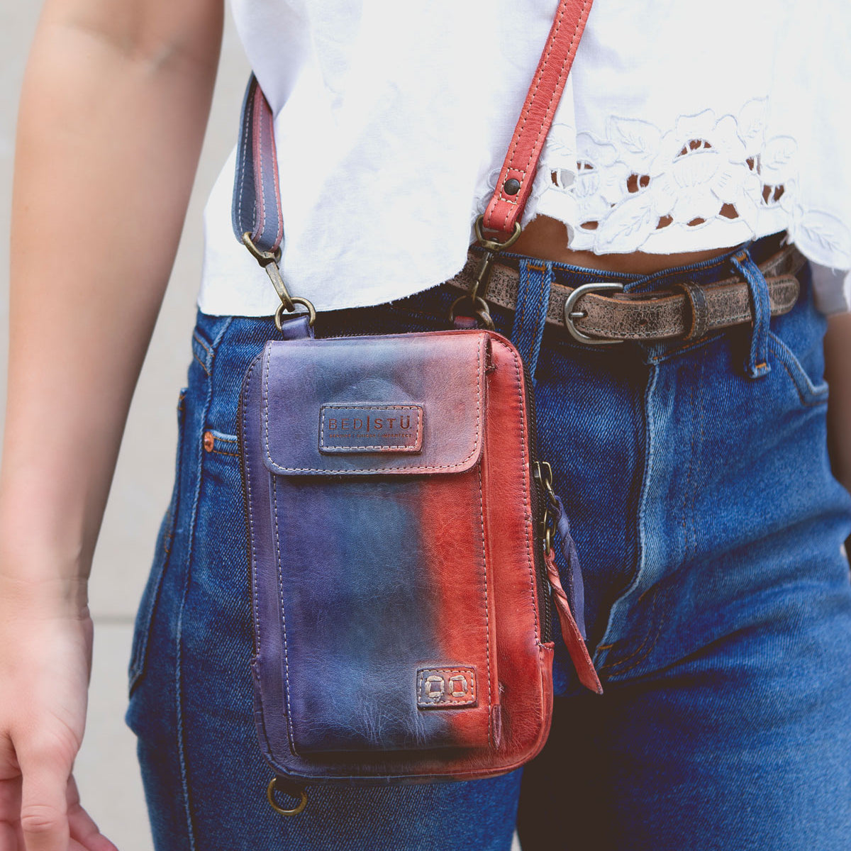 Person wearing a white top and blue jeans, carrying a small red and blue Alelike crossbody phone holder with a premium leather strap for added daily functionality from Bed Stu.