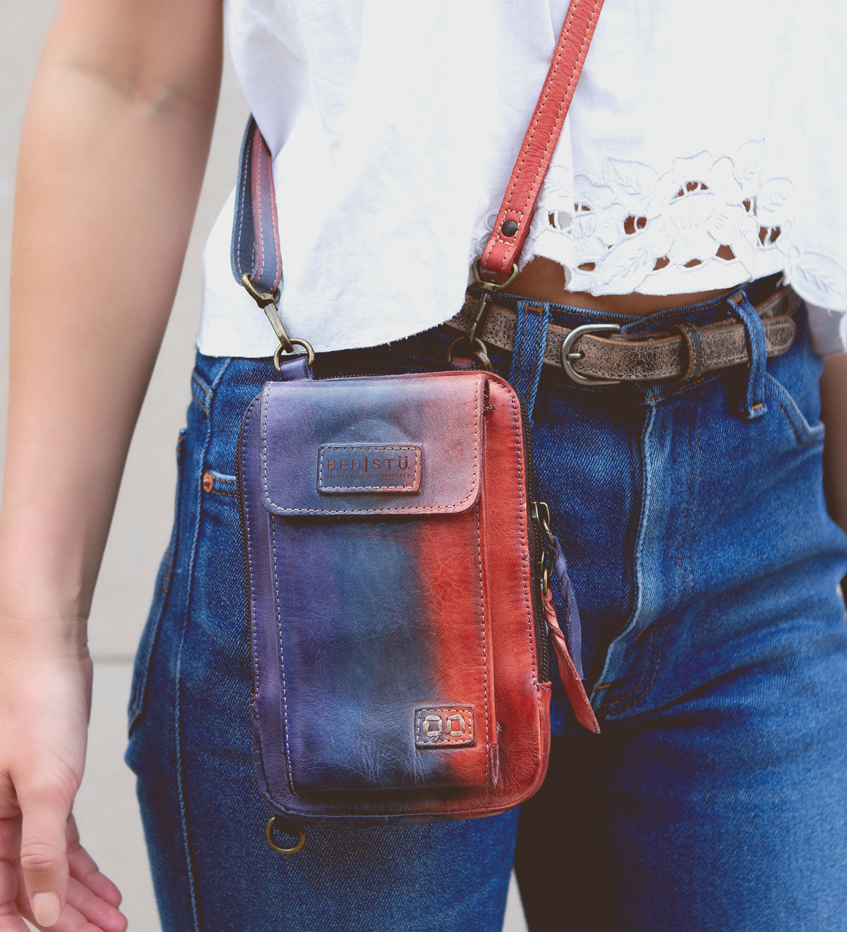 Person wearing a white top and blue jeans, carrying a small red and blue Alelike crossbody phone holder with a premium leather strap for added daily functionality from Bed Stu.