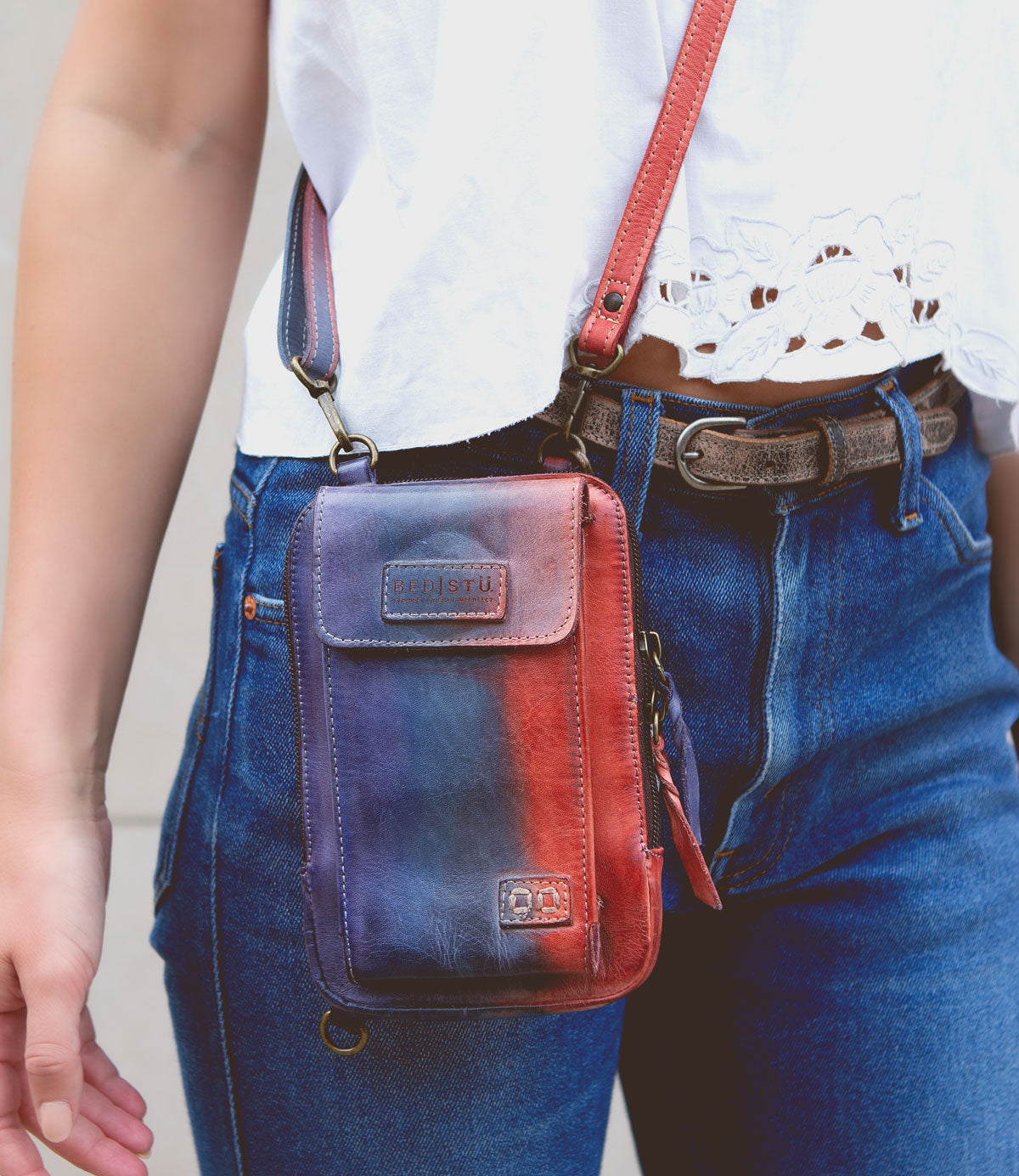 Person wearing a white top and blue jeans, carrying a small red and blue Alelike crossbody phone holder with a premium leather strap for added daily functionality from Bed Stu.