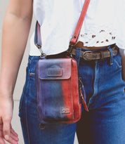 Person wearing a white top and blue jeans, carrying a small red and blue Alelike crossbody phone holder with a premium leather strap for added daily functionality from Bed Stu.
