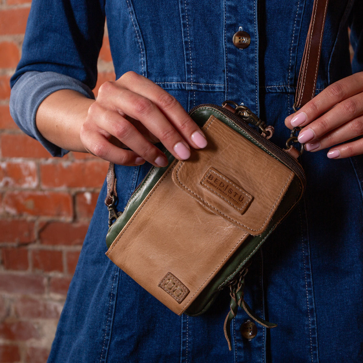 A person in a denim dress holds the Bed Stu Alelike, a premium leather compact crossbody phone holder, in front of a brick wall.